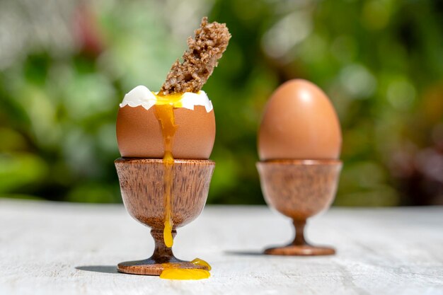 Soft boiled egg in eggcup with slice of toasted bread on white wooden table in nature background closeup