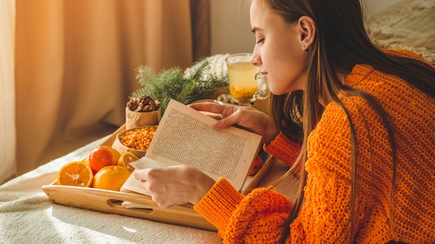 Foto accogliente e morbida di donna in caldo maglione arancione sul letto con una tazza di tè e libro.