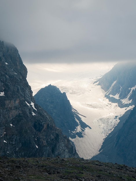 Soft focus atmospheric vertical landscape with fuzzy\
silhouettes of sharp rocks and snowy mountaintop in low clouds\
during rain dramatic view to large glacier blurred in rain haze in\
gray low clouds