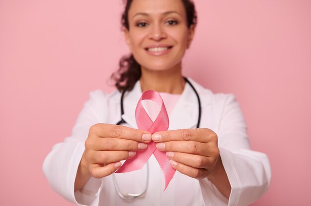 Soft focus sul nastro rosa per la consapevolezza del cancro al seno, nelle mani di una dottoressa offuscata di razza mista in camice bianco, isolato su sfondo colorato. 1 ottobre, giornata mondiale contro il cancro al seno