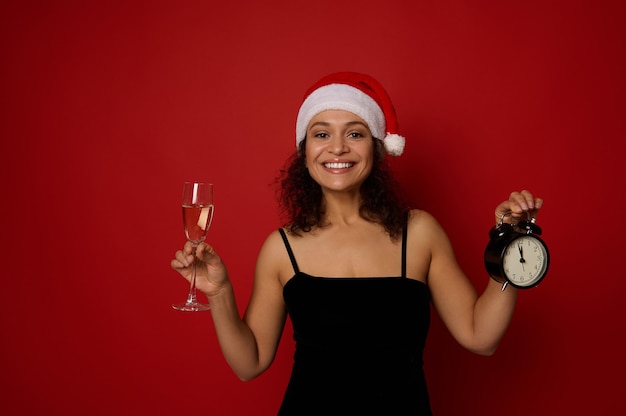 スパークリングワインと目覚まし時計を備えたシャンパンフルートに、美しい歯を見せる笑顔で笑顔のサンタ帽子をかぶったきれいな女性の手にソフトフォーカス。コピースペースと赤い背景のクリスマスの概念