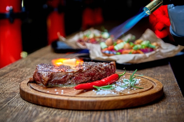 Soft focus. Entrecote Beef Grilled steak meat with fire flames on wooden cutting boardon with rosemary branch, pepper and salt . Master chef cooking delicious grill barbecue. Melt butter