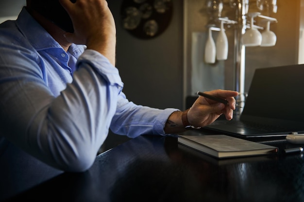 Soft focus on the hand holding a pen of a busy unrecognizable businessman, freelancer talking on mobile phone while working remotely on laptop at home office