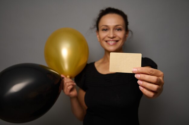 Soft focus ocus on empty blank golden credit card in the hand\
of beautiful smiling woman holding black gold air balloons, posing\
against gray wall background with copy ad space. black friday\
concept