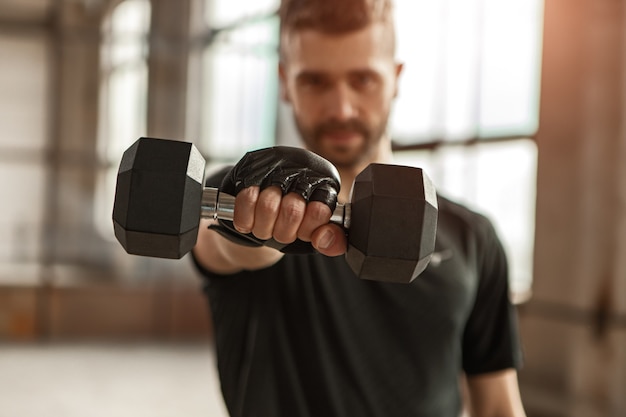 Soft focus of sportsman demonstrating heavy dumbbell to camera during fitness workout in gym