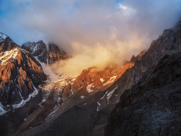 Soft focus. Sunlight in the mountains. Big glacier on top in orange light. Scenic mountain landscape with great snowy mountain range lit by dawn sun among low clouds.