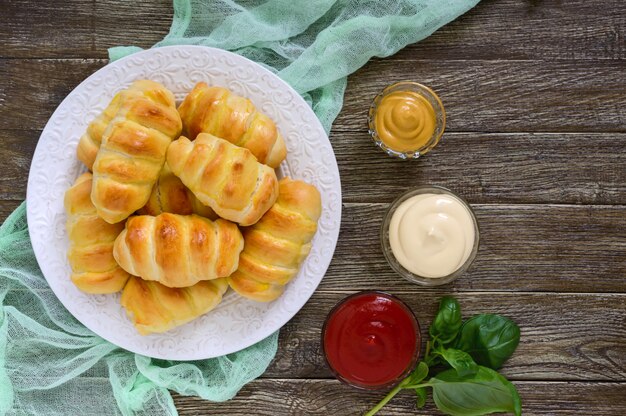 Soft homemade buns with sausage on a white ceramic plate with different sauces. Sausage in the dough. Top view.