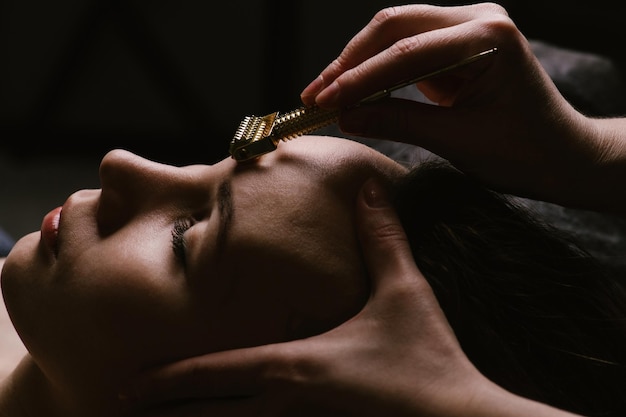 Soft light image of a woman receiving forehead massage in a beauty salon Her eyes closed She's lying on a massage bed enjoying the sensation Hands massaging her forehead with a tipped roller