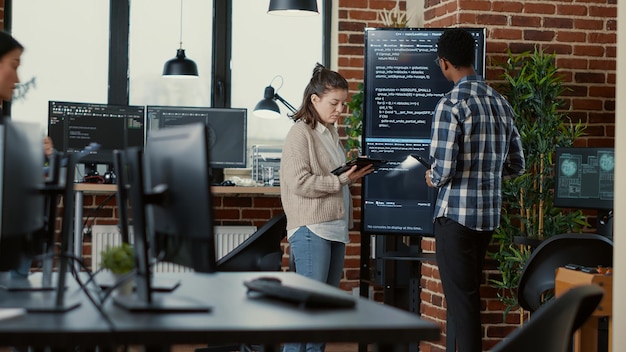 Software developer holding digital tablet analyzing code on wall screen tv explaining errors to coworker programer in busy office. Team of coders collaborating on ai cyber security group project.