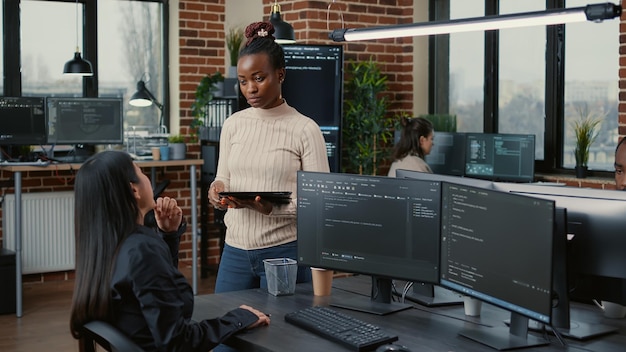 Software developer standing up holding digital tablet talking\
with programer sitting at desk with computer screens showing\
running code. system engineer coding with team writing machine\
learning data.