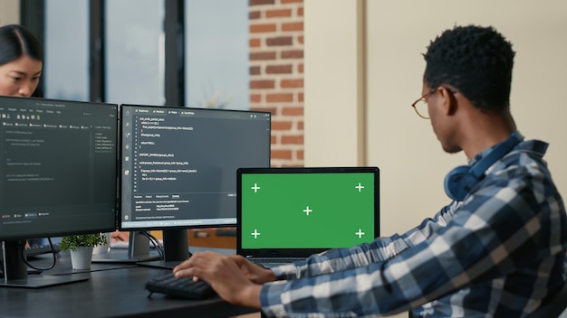 Software developer writing code using computer keyboard and laptop with green screen chroma key mockup sitting at desk. Programer looking at multiple screens with machine learning algorithm.