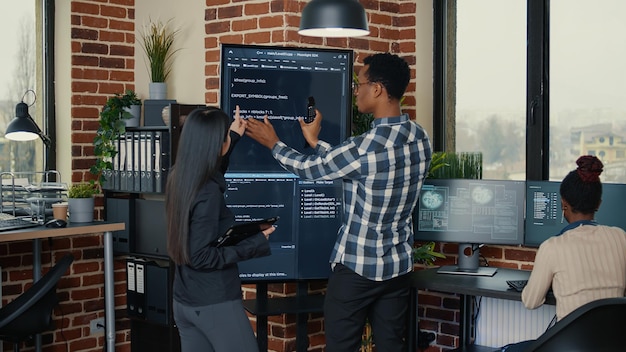 Software engineers analyzing code on wall screen tv comparing\
errors using digital tablet next to programer working with\
artficial intelligence. team of coders working on cloud computing\
project.