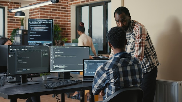 Softwareontwikkelaar die code schrijft op laptop en computertoetsenbord en naar meerdere schermen met programmeertaal kijkt, wordt onderbroken door een collega die om advies vraagt. Cloud programmeurs doen teamwerk.