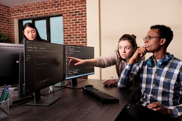 Softwareontwikkelaar vraagt coder om hulp met html-code, wijzend op database script op terminal venster. Team van programmeurs ingenieurs werken aan app ontwikkelen programma in data room.