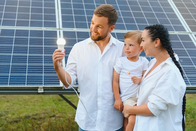 Foto il concetto di energia solare una giovane famiglia felice è in piedi vicino ai pannelli solari e tiene in mano una lampadina elettrica