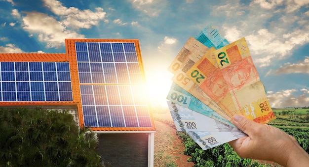 Economia dell'energia solare, con denaro brasiliano davanti al pannello fotovoltaico su un tetto, sfondo al tramonto. spazio per il testo.