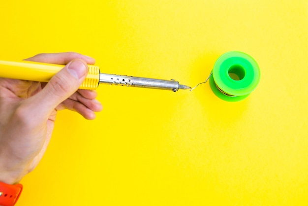 Soldering iron, tin, rosin on a yellow table. Soldering iron in male hands. Repair of electrical equipment, radio engineering. Solder wires, contacts.