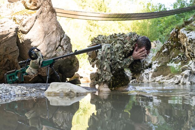 Soldato in uniforme da camuffamento che beve acqua fresca dal fiume fucile da cecchino militare sul lato