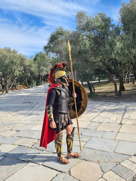 A soldier in a greek costume stands in a park with a large stone path.