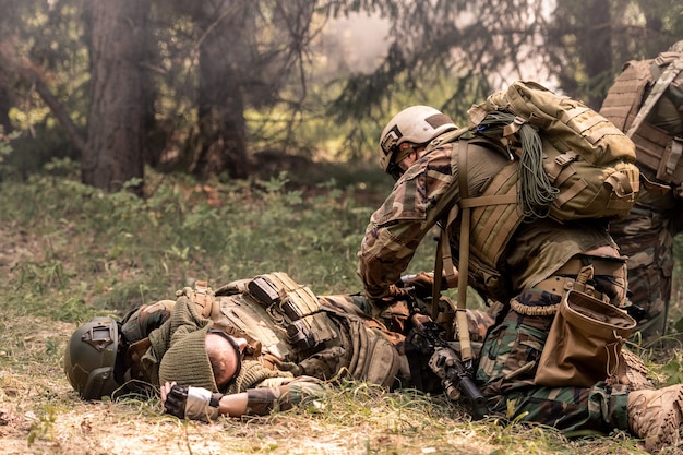 Soldier in helmet standing on knees near dead colleague and shooting on battlefield, fog in forest