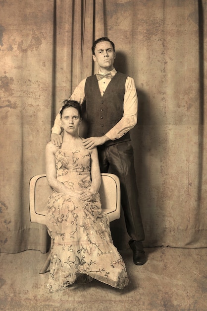 Foto solenne ritratto del matrimonio. la donna, seduta in un elegante abito a fiori. l'uomo, in piedi, in abito elegante. entrambi con espressioni serie e solenni. vecchio effetto foto