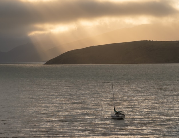 одинокая лодка, плывущая на водной глади во время восхода солнца с лучами света на фоне новой зеландии