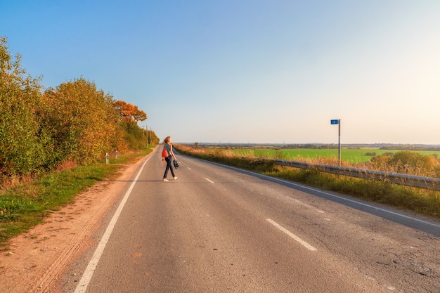 Concetto di viaggio in solitaria. il punto di vista posteriore della donna con lo zaino attraversa la strada campestre di autunno, escursione