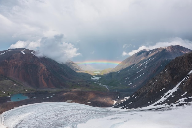 Somber landschap met levendige regenboog boven bergmeer Somber landschap met heldere regenboog boven gletsjer en gletsjermeer in bergdal Bovenaanzicht op kleurrijke regenboog en lage wolken in bergen