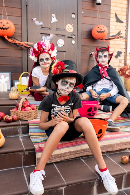 Sombere jongen met geschilderd gezicht zittend op trap voor camera tegen zijn vrienden in kostuums van halloween