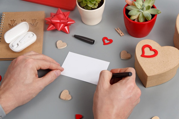 Somebody writing a card near a gift box and hearts on grey table close up