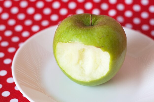 Someone bit a green apple lying on a white plate on a red polka dot background