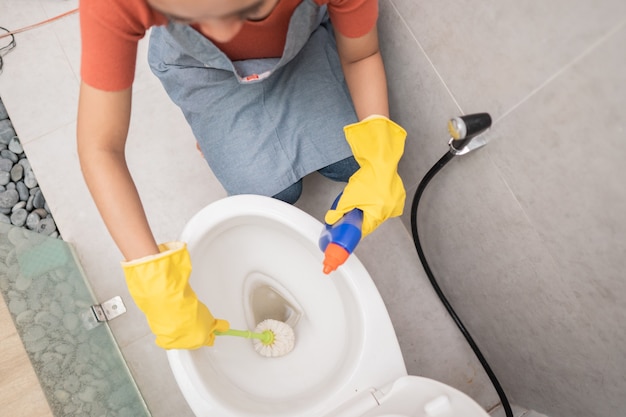 Someone wearing gloves clean the toilet with a brush and porcelain cleaner in the bathroom