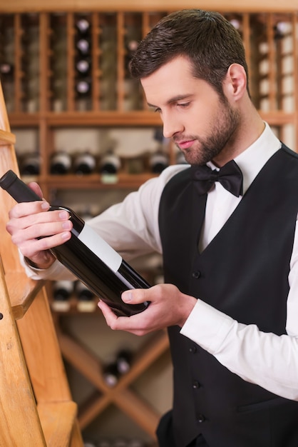 Sommelier choosing wine. Confident young man in waistcoat and bow tie holding bottle with wine and looking at it while standing in liquor store