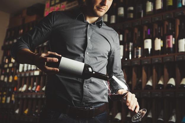 Sommelier male pouring red wine from bottle having label into glass on angle alcoholic beverage degustation cellar with containers and drinks set