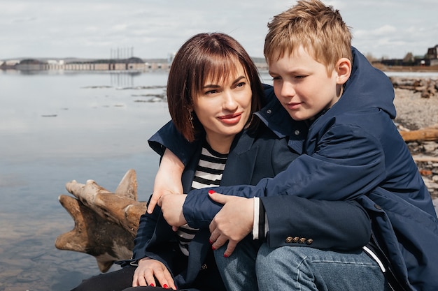 Photo a son embraces his mother on the riverbank. a beautiful, modern mother with a happy son sitting on logs.