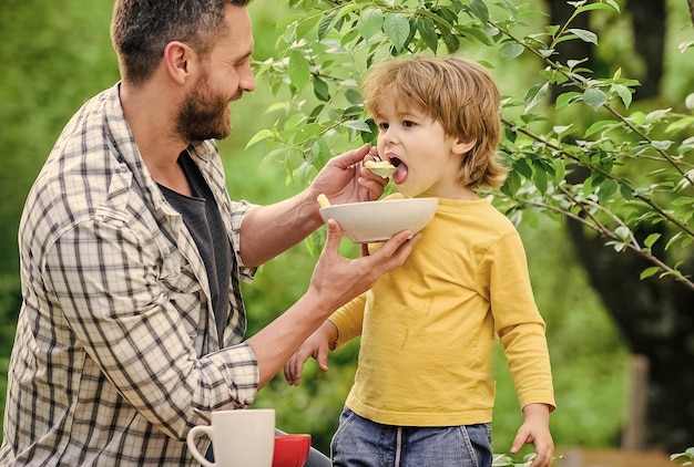 屋外で食べる息子と父親朝の朝食おいしい食べ物健康的な食べ物とダイエット乳製品幸せな父親の日お父さんと一緒の小さな男の子は穀物を食べる家族の休憩時間家族との週末