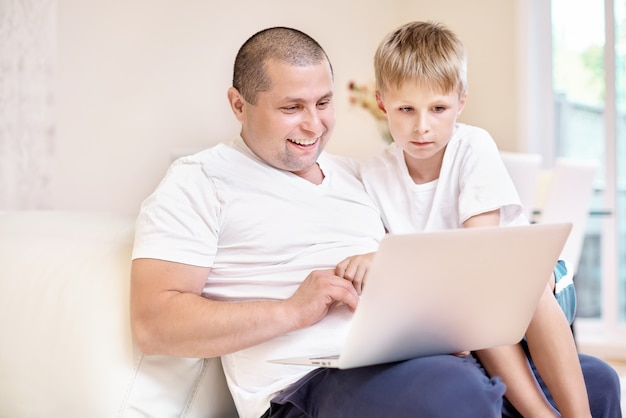 Сын и его отец сидят на диване, глядя на ноутбук, счастливые эмоции от увиденного, счастливая семья