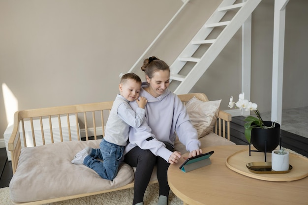 The son hugs his mom and watches her mother working on a tablet.