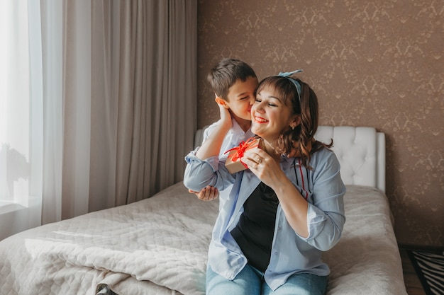 Son makes mom a present for mother's day. mom and baby sit on the bed with a gift box.