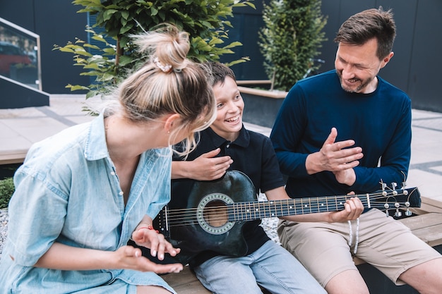 Son shows his parents how he learns to play t guitar . They sit in city square and and are happy together. Lifestyle. High quality photo