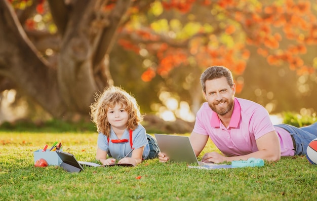 Son with father learning outdoor by studying online and working on laptop in green field