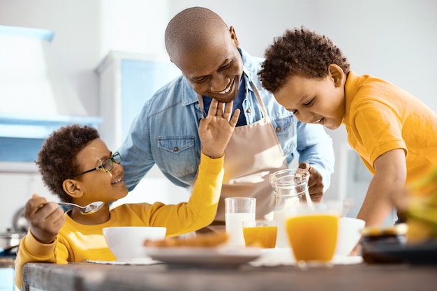 Sonly affection. Pleasant little boy caressing his fathers chin and smiling fondly while he serving his children breakfast