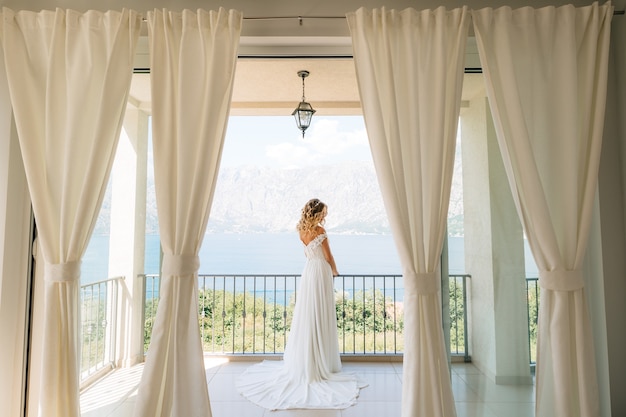 A sophisticated bride in an elegant wedding dress stands on a wide balcony with a picturesque view