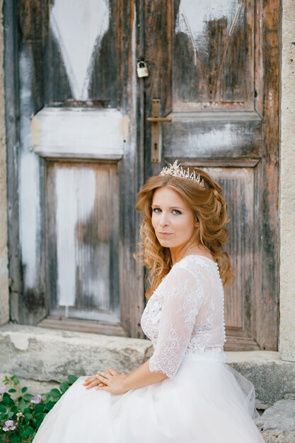 Una sposa sofisticata con tiara seduto vicino a una vecchia porta di legno nel centro storico di perast closeup