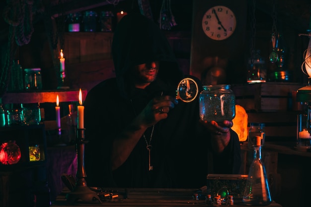 Sorcerer alchemist in dark clothes is engaged in potion making in a craft workshop with a colorful neon light