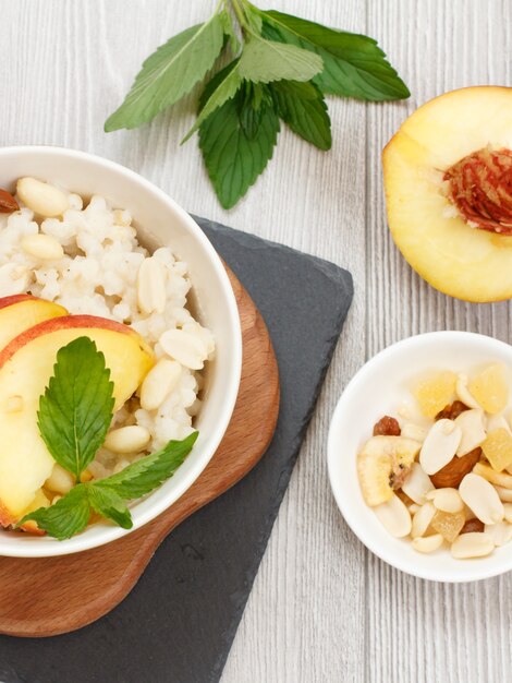 Sorghum pap met stukjes perzik, cashewnoten en amandel in porseleinen kommen, verse perziken op houten en stenen planken. Vegan glutenvrije sorghumsalade met fruit. Bovenaanzicht.