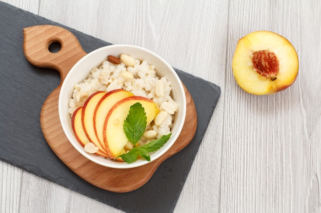 Sorghum porridge with pieces of peach, cashew nuts and almond in porcelain bowl, fresh peach on wooden and stone boards. Vegan gluten-free sorghum salad with fruits. Top view.