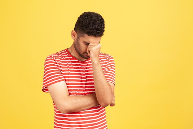 Sorrowful hopeless man in striped t-shirt lowering his head hiding eyes, upset depressed about mistake, become broke, feeling shame. Indoor studio shot isolated on yellow background