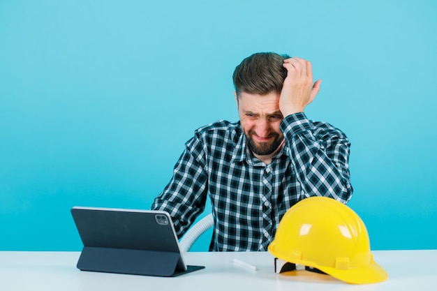 Sorry architect is holding hand on head by sitting in front of tablet on blue background