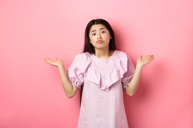 Sorry cant help. Clueless asian girl shrugging with sad face, know nothing, standing indecisive against pink background.